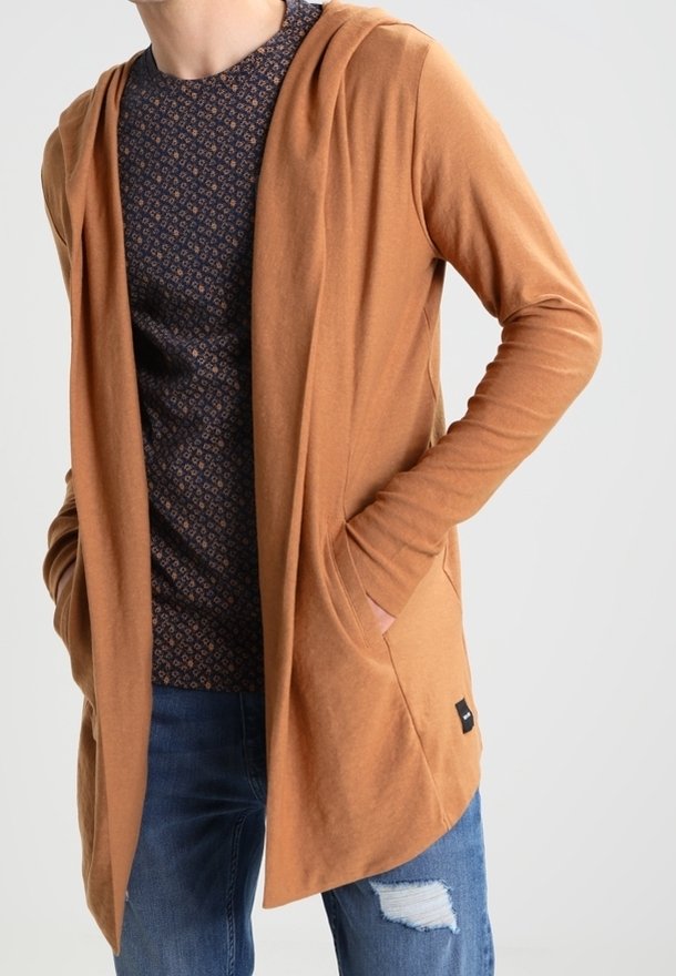 Personne portant un sweat à capuche marron clair ouvert, une chemise foncée à motifs et un jean bleu déchiré, les mains dans les poches du sweat.