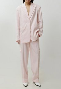 Personne portant un costume rose pâle surdimensionné avec des poches cargo, associé à des bottes blanches à bout pointu, se tenant devant un fond uni.