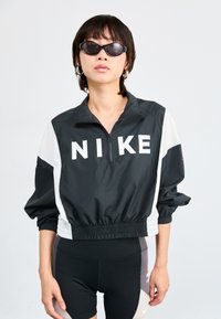 Coupe-vent Nike noir et blanc à zip quart, avec des poignets et un ourlet élastiques, associé à un short de cyclisme noir.