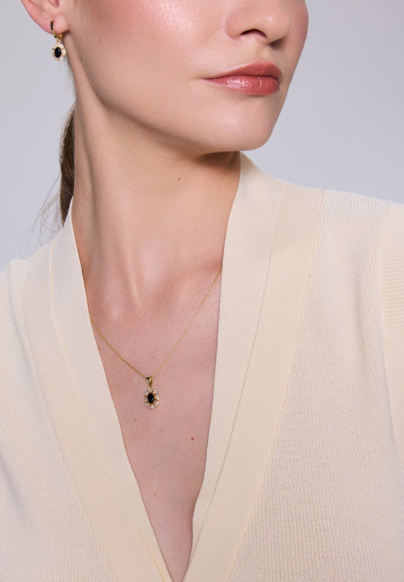 Złoty naszyjnik z czarnym wisiorkiem, otoczony przez przezroczyste kamienie. Modelka ma na sobie kremowy sweter z prążkowanego materiału z dekoltem w kształcie litery V i gładką teksturą.