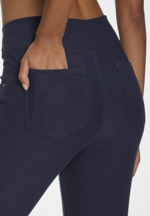 Hand in der hinteren Tasche einer eng anliegenden navyblauen Hose mit hohem Bund, zeigt die Stoffstruktur und das Detail der Taschennaht.