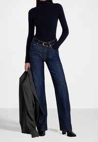 Haut à col roulé côtelé bleu marine, jeans évasés en denim foncé taille haute, ceinture en cuir noir avec boucle en or, blazer gris drapé sur le bras.
