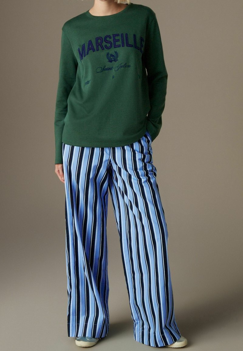 Grüner Pullover mit dem Schriftzug "MARSEILLE" in Dunkelblau, kombiniert mit weit geschnittenen blauen und weißen gestreiften Hosen. Lässiger, lockerer Schnitt und Textur.