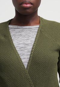 Vihreä neulottu neulejakku, jossa on V-pääntie, teksturoitu kangas ja ribbireunat, käytetty harmaan raidallisen pitkähihaisen paidan päällä.