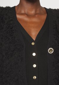 Schwarze Strickjacke mit flauschiger Textur, V-Ausschnitt, goldenen Knöpfen und einer dekorativen Brosche mit kreisförmigem Design und Strasssteinen.