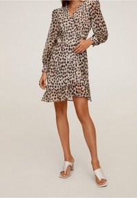 Femme portant une robe portefeuille à imprimé léopard, à manches longues et à mi-mollet, avec des sandales à talons ouvertes blanches, se tenant devant un fond uni.