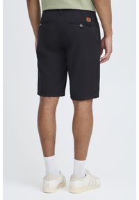 Svarte shorts med en glatt tekstur, to baklommer med knapper, og en lys lærlapp på livet. Brukt med hvite sneakers.