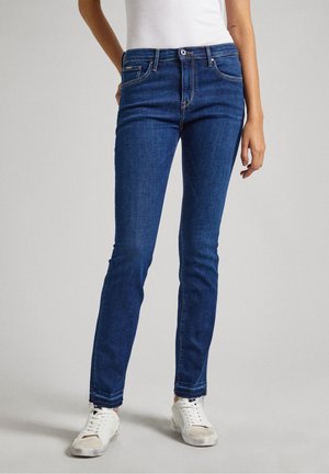 Vrouw draagt blauwe slim-fit jeans met rafelige zomen en witte sneakers, staand tegen een effen lichte achtergrond.