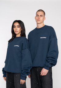 Felpe blu navy con logo "MULTIPLY" in bianco, vestibilità ampia, collo a giro ribattuto e polsini, indossate con jeans scuri, tessuto testurizzato.