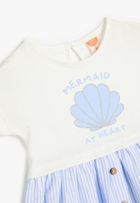 Valkoinen paita, jossa on sininen kuori -grafiikka ja teksti "MERMAID AT HEART"; siniraitainen hame, jossa on puunapit. Pehmeä puuvillamateriaali.