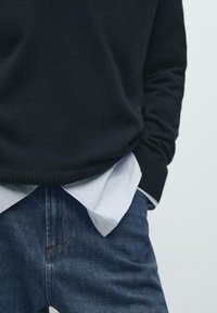 Czarny dziergany sweter z prążkowanym dołem na jasnoniebieskiej koszuli z kołnierzykiem, zestawiony z ciemnoniebieskimi dżinsami. Zbliżenie na dolną część tułowia.