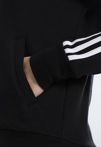 Sweat-shirt noir avec une poche kangourou, présentant trois bandes blanches le long du poignet. Le tissu semble doux et texturé.