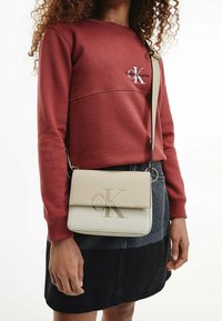 Sac bandoulière beige avec logo "CK" embossé, texture en cuir lisse, accessoires argentés, assorti à un sweat-shirt bordeaux et une jupe en denim.