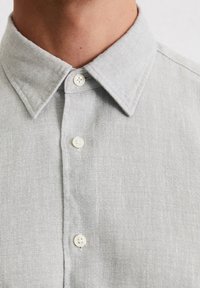 Chemise à boutons gris clair en tissu texturé, avec un col classique et des boutons blancs. Le design présente une coupe légèrement décontractée.