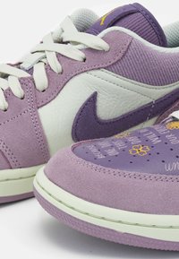 Baskets Nike violettes et blanches avec textures en daim et cuir, lacets blancs, swoosh violet et texte décoratif en forme de fleur jaune sur la boîte à orteils.