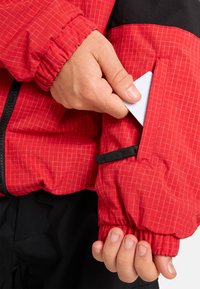 Rote Jacke mit Karomuster und schwarzem Akzent. Der Ärmel hat eine enge Manschette und eine Tasche, in die teilweise eine weiße Karte gesteckt ist.