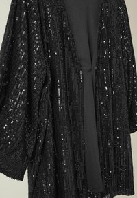 Châle en sequins noirs avec une coupe décontractée, comportant une couche intérieure noire lisse et unie. Des sequins scintillants créent une surface texturée et réfléchissante.