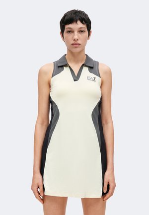 Mujer con cabello corto y oscuro, vistiendo un vestido ajustado sin mangas en crema y negro con cuello y logo EA7 en el pecho, de pie frente a un fondo liso.