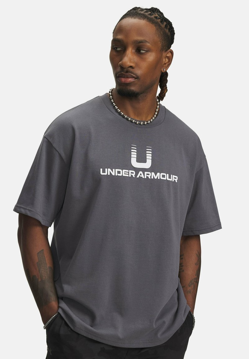 Grå bomulds-t-shirt med rund hals, korte ærmer og et stort hvidt "UNDER ARMOUR" logo på forsiden. Glat tekstur.
