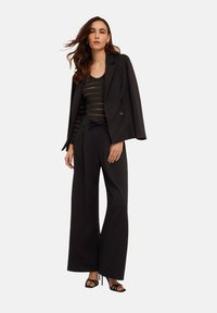 Svart skräddarsydd kavaj med dubbla knappar, lager på lager över en randig svart topp och vida svarta byxor. Outfiten har mjuka texturer och minimala accenter.