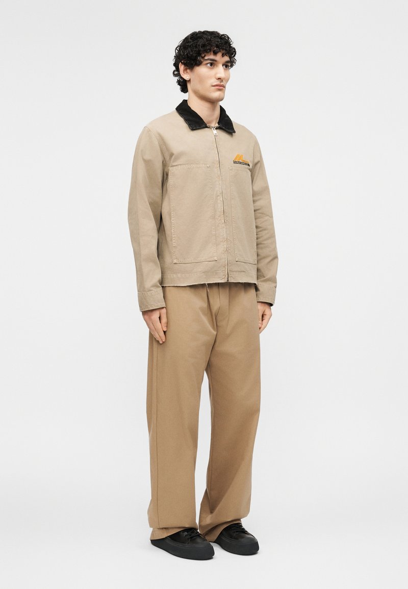 Jonge man staat frontaal gekeerd en draagt een beige jas met zwarte kraag, beige broek met wijde pijpen en zwarte sneakers op een witte achtergrond.