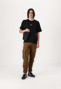 Schwarzes T-Shirt mit kurzen Ärmeln und Logo, braune Cordhose mit Taschen und schwarze Sneakers. Kleine schwarze Umhängetasche.