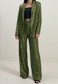 Groene pailletten suit met een getailleerde jas en broek met wijde pijpen. De stof heeft een glanzende textuur, gecombineerd met een zwarte cropped top en zwarte hakken.