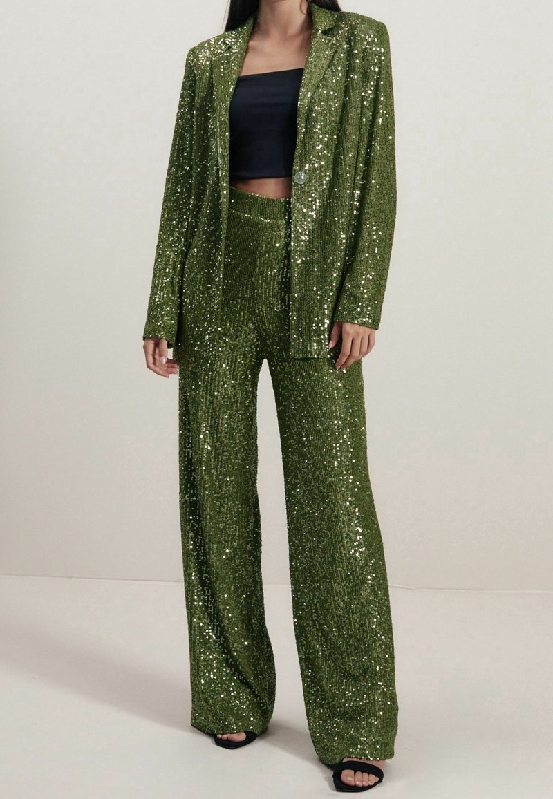Groene pailletten suit met een getailleerde jas en broek met wijde pijpen. De stof heeft een glanzende textuur, gecombineerd met een zwarte cropped top en zwarte hakken.