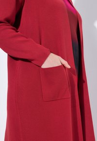 Rode cardigan met lange mouwen, geribbelde textuur en een patchzak aan de linkerkant. De stof lijkt zacht en lichtgewicht.