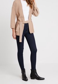 Beige stickad cardigan med vida ärmar och knytbälte i midjan, bärs över en vit t-shirt, tillsammans med mörkblå skinny jeans och svarta ankelboots.