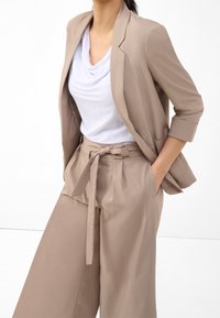 Femme portant un blazer beige et un pantalon large avec une ceinture à nouer, associé à un top blanc à encolure drapée, les mains dans les poches.