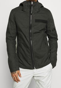 Mörkgrön zip-up jacka med huva, utrustad med en bröstficka, elastiska ärmslut och en slät tygkänsla. Accentuerad med svarta detaljer.