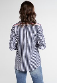 Navy and white striped shirt with a back pleat, roll-up sleeves, and a red accent on the collar. Made from a smooth fabric.