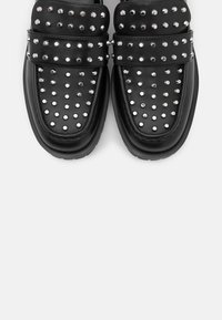 Mocassins en cuir noir avec des clous ronds argentés couvrant le dessus et un détail de sangle sur le cou-de-pied, vus de dessus.