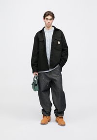 Czarna kurtka zapinana na guziki, jasnoszary sweter w warkocze, luźne ciemne dżinsy, beżowe buty, trzymająca małą torbę w kolorze morskim.