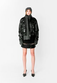 Chaqueta bomber de cuero negro con múltiples bolsillos, combinada con una falda negra ajustada adornada con cierres metálicos. Bufanda y gorro de punto gris.