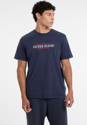 Granatowa bawełniana koszulka z krótkim rękawem, z białym i czerwonym logo graficznym z napisem "GUESS JEANS" na środku przodu.