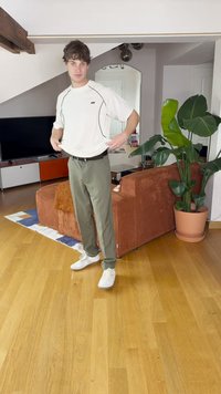 Λευκό αθλητικό πουκάμισο με μαύρες λεπτομέρειες, λάδι πράσινα παντελόνια και λευκά αθλητικά παπούτσια. Στέκεται σε ένα σαλόνι με ένα φυτό και έναν καναπέ.