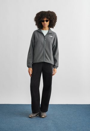 Ženska z kodrastimi lasmi, ki nosi črna sončna očala, sivo jakno na zadrgo iz flisa, črne hlače in sive superge, stoji na modrem preprogi.