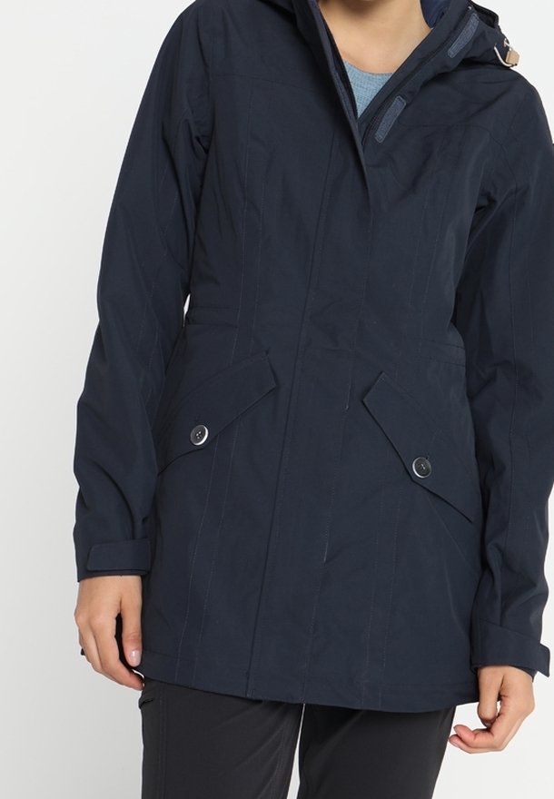 Femme portant une veste à capuche bleu marine avec des poches à rabat boutonnées et une fermeture éclair frontale, associée à un pantalon noir.