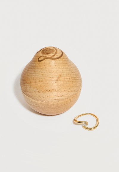 Træbeholder med afrundede kanter og udskåret swirl-design ved siden af en åben guldring med en lille klar sten, på en hvid overflade.