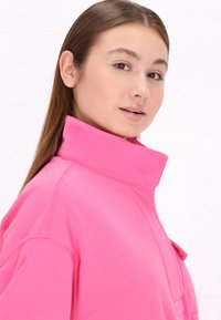 Felpa rosa con colletto alto, texture morbida e tasca frontale. Il design include dettagli di cucitura e una vestibilità comoda.