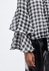 Μπλούζα με ασπρόμαυρο καρό, με μανίκια καμπάνα από ελαφρύ ύφασμα, συνδυασμένη με γυαλιστερό μαύρο δερμάτινο παντελόνι.