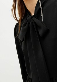 Frau tr ägt eine schwarze Bluse mit einer großen Schleife am Kragen und einem langen, hängenden Strassohrring auf einer Seite.