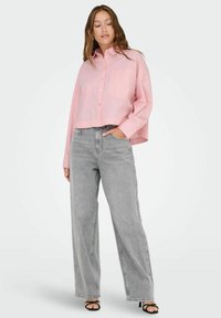 Camicetta rosa corta con design a bottoni e tasca sul petto, abbinata a jeans a gamba larga grigi a vita alta e sandali neri con tacco.