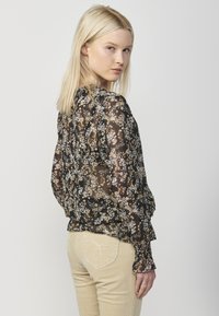 Blusa nera trasparente con motivi floreali in crema e senape, polsini arricciati e una silhouette leggermente affusolata, abbinata a pantaloni in corda beige chiaro.