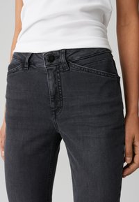 Jean en denim noir avec une coupe slim, présentant une fermeture à bouton, des passants de ceinture et des poches avant. Le tissu a une texture lisse avec un léger délavage.