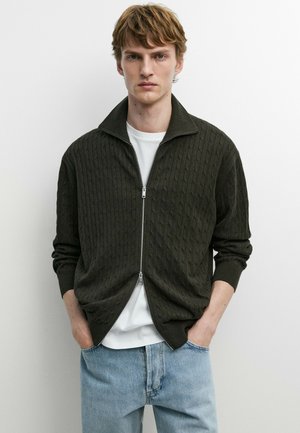 Junger Mann mit hellbraunen Haaren, trägt einen dunkelgrünen grob gestrickten Reißverschluss-Pullover über einem weißen T-Shirt und hellblauen Jeans, steht mit Händen in den Taschen.