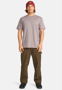 Šedé tričko s krátkým rukávem s logem na hrudi, doplněné volnými hnědými cargo kalhotami a černými teniskami, doplněné červenou čepicí.