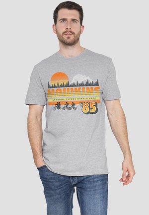 Gray cotton t-shirt featuring a multicolored graphic with a sun, mountains, forests, and bicycles. Text states "HAWKINS" and "Strange Things Happen Here '85."
